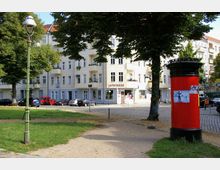 Ein kleiner städtischer Park mit einem Weg und Rasenflächen, umgeben von mehrstöckigen Wohnhäusern mit hellfarbenen Fassaden. Im Vordergrund stehen eine grüne Altstadtlaterne und eine rote Litfaßsäule, im Hintergrund sind geparkte Autos und eine Apotheke zu sehen.