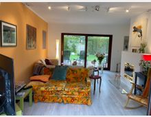 Wohnzimmer mit großen Glastüren, die zu einem Garten führen. Der Raum ist mit farbenfrohen Möbeln, einer gelb-orange gemusterten Couch, einem modernen Kamin und dekorativen Kunstwerken an den Wänden eingerichtet.