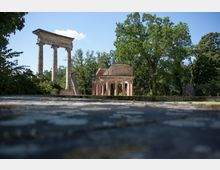 Ruinen eines antiken Tempels mit zwei erhaltenen Säulen und einer halbrunden Kolonnade aus Backstein, umgeben von dichtem Grün und Bäumen unter klarem Himmel.