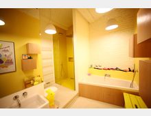 Modernes Badezimmer mit gelben Wänden, einer weißen Waschbeckenarmatur, einer Badewanne mit Holzelementen, einer begehbaren Dusche mit Glaswand und einer weißen Backsteinwand. Ein gelber Hocker und spielerische Deko-Elemente verleihen dem Raum eine kindliche Note.