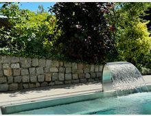 Swimmingpool mit integriertem Wasserfall aus einer gebogenen Metallkonstruktion, umgeben von einer Steinmauer und dichter, grüner Gartengestaltung mit Büschen und Bäumen. Im Hintergrund ist ein sonniger Himmel sichtbar.