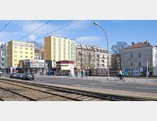 Straßenszene mit gelbem Hotelgebäude, mehrstöckigen Wohnhäusern und einer Sparkassen-Filiale im Vordergrund. Straßenbahnschienen, Fußgänger und ein Radfahrer prägen die belebte Umgebung, während Werbeplakate eine nahegelegene Mauer bedecken.