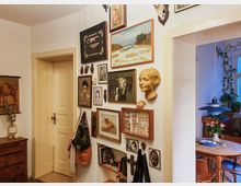 Ein Innenraum mit einer weißen Wand, die mit diversen Bildern, Gemälden und Dekorationen (wie einem Wandrelief einer Frauenbüste und Insektenrahmen) geschmückt ist. Links eine geschlossene, cremefarbene Tür und rechts ein Durchgang zu einem Esszimmer mit Holztisch, Stühlen und Zimmerpflanzen.