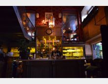 Gemütliche Bar mit dunkler Holz-Theke und einer Wand voller beleuchteter Flaschen und Gläser. Im Hintergrund sind eine Wendeltreppe, rote Wände mit Mustertapeten und ein Kronleuchter zu sehen.