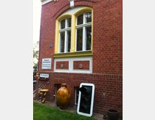 Backsteinfassade eines Gebäudes mit gelb umrahmten Rundbogenfenstern. Im unteren Bereich befinden sich Dekorationsobjekte wie eine große Keramikvase und ein umgestürztes Verkehrssignal, sowie ein Schild mit der Aufschrift „Richardplatz“.