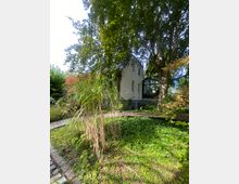 Ein Garten mit dichter Vegetation, darunter hohe Gräser, Bäume und Büsche, umgibt ein zweistöckiges Haus mit heller Fassade. Ein hölzerner Weg verläuft geschwungen durch die grüne Fläche.