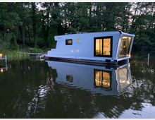 Ein modernes, weißes Hausboot mit großen Fenstern liegt auf einem ruhigen See, umgeben von dichten, bewaldeten Ufern. Im Inneren des Hausbootes ist warmes Licht zu sehen, das den Innenraum erhellt.