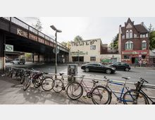 Straßenszene mit einer Brücke aus Stahl, auf der ein Graffito mit der Aufschrift "KEINE RINDERZUCHT" zu sehen ist, darunter der S-Bahnhof "Yorckstraße". Im Vordergrund sind mehrere Fahrräder an einem Geländer abgestellt, daneben fahren Autos, und im Hintergrund steht ein Backsteingebäude mit kleinen Geschäften.