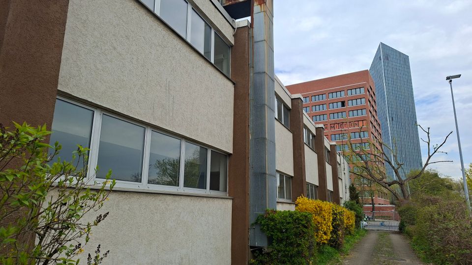 Schmaler Gehweg entlang eines flachen Büro- oder Verwaltungsgebäudes mit heller Putzfassade, langen Fensterbändern und einem senkrechten Metallschacht. Im Hintergrund stehen ein rotes Hochhaus und ein schlanker gläserner Büroturm, umgeben von Sträuchern und Bäumen.