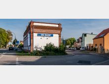 Eckgebäude mit Backsteinfassade und einem Schild mit der Aufschrift „US-Shop“, umgeben von niedrig bewachsenen Pflanzen. Im Hintergrund eine Wohnstraße mit farbigen Häusern, geparkten Autos und vereinzelten Bäumen entlang der Straße.