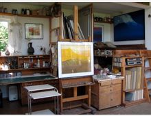 Ein Künstleratelier mit einer Staffelei in der Mitte, auf der ein Kunstwerk steht. Im Hintergrund sind Regale mit Farben, Pinsel, Bücher, eine Skulptur und Dekorationsgegenstände zu sehen, sowie ein Hochbett und weitere Möbel mit Kunstmaterialien.