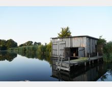 Holzhütte am Ufer eines stillen Gewässers mit angrenzendem Holzsteg und Leiter, umgeben von Schilf und Bäumen, die sich im klaren Wasser spiegeln. Der Himmel ist wolkenlos und hellblau.