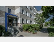 Mehrstöckiges Wohn- oder Bürogebäude mit einer grauen Fassade aus grobem Putz und weißen Fensterrahmen. Der Eingangsbereich verfügt über eine blaue Umrahmung, eine Treppe mit Geländer und ist von gepflegten Büschen und einem gepflasterten Gehweg umgeben.