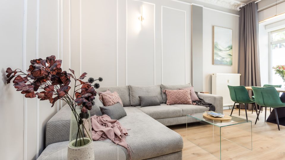 Helles, modernes Wohnzimmer mit großem grauen Ecksofa, Zierleisten an der Wand und einem gläsernen Couchtisch auf hellem Holzboden. Rechts schließt ein Essbereich mit grünen Stühlen an, neben bodentiefen Vorhängen und einem großen Fenster.