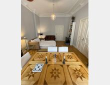 Ein Wohnraum mit einem Holztisch im Vordergrund, verziert mit sternförmigen Intarsienmustern. Im Hintergrund sind ein Sofa, ein Bett, ein Bücherregal und eine Zimmertür zu sehen, umgeben von hohen, hellgrauen Wänden und einer weißen Stuckdecke.