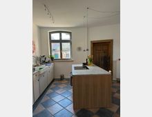 Helle Wohnküche mit großem Sprossenfenster, weißen Einbauschränken entlang der linken Wand und einer zentralen Kücheninsel aus Holz mit heller Arbeitsplatte und Kochfeld. Sichtbare Pendelleuchten an Kabeln, eine dunkle Holztür rechts und ein diagonal verlegter Fliesenboden in Blau- und Rosatönen.
