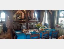 Ein rustikaler Gastraum mit Holzbalkenkonstruktion, in dessen Mittelpunkt eine Bar mit Hockern steht. Ein großer Esstisch mit blauer Tischdecke ist für eine Mahlzeit gedeckt, geschmückt mit Tulpen und verschiedenen Speisen.