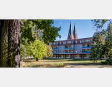 Wohngebäude mit vier Stockwerken, blauen Balkonen und rotem Dach, umgeben von Bäumen und einer Grünfläche im Vordergrund; im Hintergrund sind die spitzen Türme einer Kirche sichtbar.