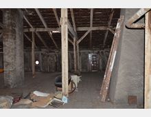 Ein leeres, verfallenes Dachgeschoss mit Holzbalkenkonstruktion und einem schrägen Dach. Zu sehen sind lose Gegenstände wie eine Leiter, ein Stuhl und zerstreute Kartons sowie eine kleine runde Lichtöffnung in der Wand.