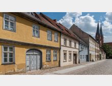 Straßenansicht einer Altstadt mit historischen Häusern in verschiedenen Farben, darunter ein gelbes Gebäude mit Holztor und kleinen Fenstern. Im Hintergrund sind eine gepflasterte Straße und eine Kirche mit hohen Türmen erkennbar.