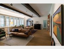 Wohnzimmer mit Essbereich in einem großzügigen Raum mit Holzbalkendecke. Im Vordergrund stehen eine braune Couch, ein Sessel und ein kleiner Tisch auf einem Teppich, im Hintergrund ein Esstisch mit Stühlen, ein Klavier, Regale und große Fenster mit dekorativen Glasdetails.