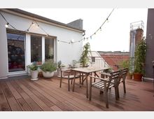 Eine moderne Dachterrasse mit Holzbohlenboden, ausgestattet mit einem Holztisch und vier Stühlen. Die Umgebung zeigt dekorative Pflanzen in Töpfen, eine Glas-Schiebetür mit Blick ins Gebäudeinnere und bunte Lichterketten, die quer über den Bereich gespannt sind.