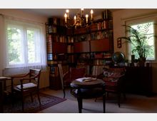 Wohnzimmer mit Bücherregal und Sitzgelegenheit. Der Raum enthält ein großes Wandregal gefüllt mit Büchern, zwei Fenster mit weißen Vorhängen, eine Sitzgruppe mit gepolsterten Sesseln und einem Tisch sowie eine Kommode mit Pflanzen und einem Globus.