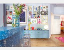 Bright, colorful interior with a speckled blue bar counter and clear stools, facing a pale blue wall covered in framed artwork above a low dresser with decorative objects and a globe lamp. A metallic pendant light hangs from the ceiling, and a doorway opens to an adjacent room with wood floors and a pink rug.