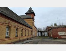 Innenhof einer historischen Backsteinanlage mit langem gelb-rotem Ziegelgebäude, Rundbogenfenstern und einem Turm mit steilem Dach. Rechts steht ein niedriger Backsteinbau; dazwischen führt ein überdachter Durchgang zu einem Tor, umgeben von grauem, rissigem Asphalt und vereinzelten Bäumen im Hintergrund.