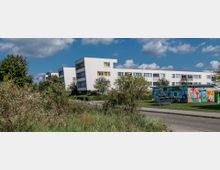 Mehrfamilienhäuser im modernen Stil mit weißen Fassaden, umgeben von Grünflächen und Gehwegen; im Vordergrund ein kleiner, bunt bemalter Pavillon mit Graffiti. Der Himmel ist leicht bewölkt, und die Umgebung wirkt städtisch und gepflegt.