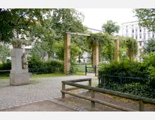 Ein kleiner Stadtpark mit einer Skulptur aus Stein, die Bären darstellt, sowie einer gelben Backsteinpergola, die mit Kletterpflanzen bewachsen ist. Im Hintergrund sind hohe Wohnhäuser und üppiges Grün zu sehen, eingefasst von Parkzäunen.