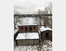Zwei verlassene Backsteingebäude mit schneebedeckten Dächern in einer winterlichen Landschaft. Im Hintergrund sind schneebedeckte Felder und kahle Bäume zu sehen, unter einem grauen, bewölkten Himmel.