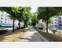 Schmaler Fußweg in einem grünen Parkstreifen, gesäumt von Bäumen, mit Wohnhäusern und Geschäften auf beiden Seiten der Straße. Im Hintergrund sind parkende Autos, Fahrräder und Menschen sichtbar.