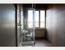 Schmales, modern gefliestes Badezimmer mit Badewanne und Glas-Duschabtrennung sowie kleinem Waschbecken und Toilette. Am Ende des Raums befindet sich ein großes Fenster mit Heizkörper darunter, das viel Tageslicht hereinlässt.