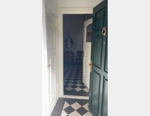 Geöffnete grüne Haustür mit goldener Türklinke, die in einen Eingangsbereich mit schwarz-weißen Schachbrettfliesen führt. Im Hintergrund sind eine Kommode, ein runder Wandspiegel und ein Stuhl sichtbar.