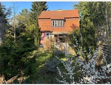 Zweistöckiges Haus mit rotem Ziegeldach, umgeben von einem üppigen Garten mit Bäumen, Sträuchern und blühenden Zweigen im Vordergrund. Im Hintergrund sind ein Holzzaun und dichte Vegetation zu sehen.