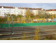 Mehrstöckige Altbauten mit hellen Fassaden im Hintergrund, davor grüne Lärmschutzwände und Bahngleise mit Oberleitungen, umgeben von herbstlicher Vegetation.