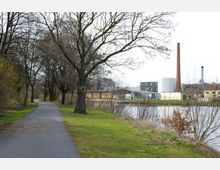 Ein asphaltierter Weg führt durch eine von Bäumen gesäumte Parklandschaft entlang eines Gewässers, mit Blick auf industrielle Gebäude und einen hohen roten Schornstein im Hintergrund. Die Szene zeigt eine Mischung aus Natur und urbaner Umgebung.