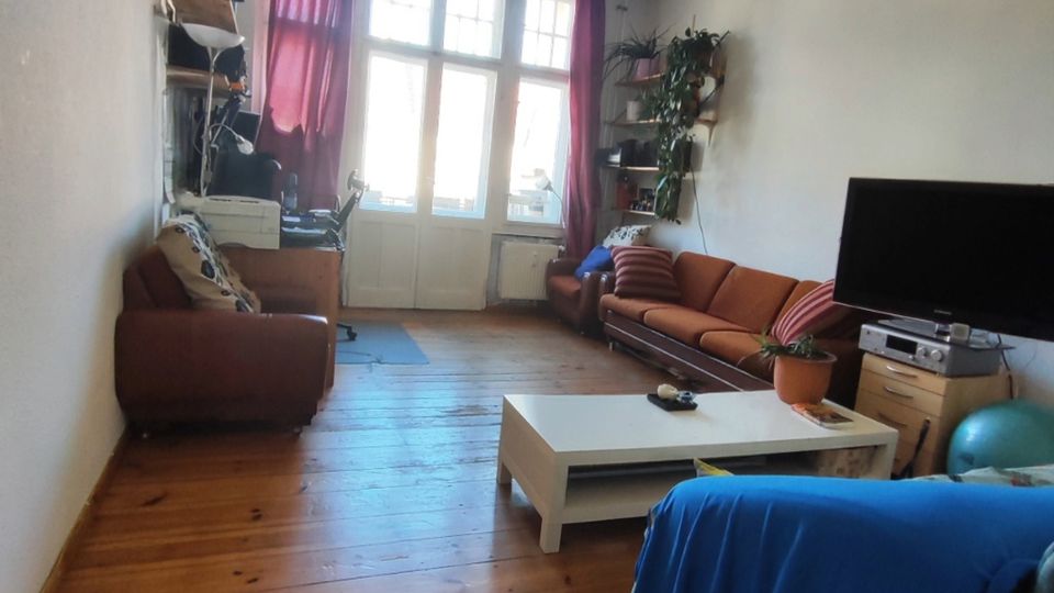 Long, narrow living room with wooden plank flooring, white walls, and a central ceiling light. Large multi-pane windows and a glass door at the far end are framed by red curtains, with sofas along the walls, a TV on a stand, and a white coffee table in the middle.