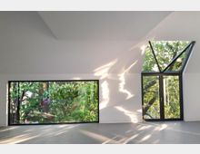 Minimalistischer, heller Innenraum mit großen Fenstern; ein rechteckiges und ein dreieckiges Fenster ermöglichen den Blick auf grüne Bäume draußen, während Sonnenlicht Muster an den weißen Wänden erzeugt.