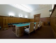 Ein holzvertäfelter Konferenzraum mit einem großen, grünen Tisch in der Mitte, umgeben von gepolsterten Stühlen. Die Wände sind mit Holzpaneelen verkleidet, die Decke ist verziert, und eine große Tür sowie moderne Wandleuchten sind sichtbar.