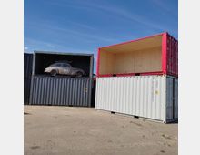 Stapel aus Frachtcontainern auf einer gepflasterten Fläche, darunter ein grauer Container mit einem Oldtimer-Auto im Inneren und daneben ein weißer und ein roter Container mit offenen Frontseiten. Der Himmel ist blau und wolkenlos.