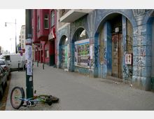 Straßenszene in einer urbanen Umgebung mit farbig verputzten Gebäuden, darunter ein rotes Haus und eine blaue, teils mit Graffiti und Plakaten bedeckte Fassade. Auf dem Gehweg liegt ein umgefallenes Fahrrad, daneben sind parkende Autos und ein Werbemast zu sehen.