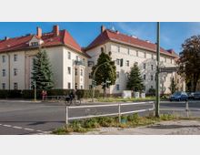 Mehrfamilienhaus aus der Vorkriegszeit mit heller Fassade, rotem Ziegeldach und kleinen Balkonen, umgeben von Bäumen und einer Hecke. Im Vordergrund eine Straßenecke mit einem Straßenschild, Fußgängern, einem Radfahrer und geparkten Autos.