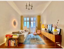 Wohnzimmer mit Holzdielenboden, einer großen cremefarbenen Couch, einem hölzernen Beistelltisch mit Büchern und einem modernen Sideboard an der rechten Wand. Gelbe Vorhänge rahmen ein Fenster mit Tür zum Balkon, und ein runder Wandspiegel mit Beleuchtung ist an der linken Wand angebracht.