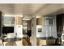 Innenraum eines modernen Wohnmobils mit Holzelementen. Sichtbar sind eine kleine Küchenzeile mit Spüle, Herdplatte und einem Wandfernseher, sowie ein schmaler Durchgang, der zu einem Schlafbereich mit Fenster führt.