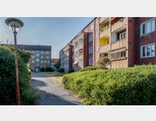 Mehrstöckige Wohngebäude mit Balkonen in einer Wohnsiedlung, umgeben von Grünflächen und Gehwegen. Im Vordergrund befindet sich eine Laterne neben gepflegten Hecken, während ein weiteres Gebäude im Hintergrund zu sehen ist.