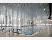 Großes Hallenbad mit einem rechteckigen Becken, umgeben von einer hohen Decke und großen Fensterfronten, die Tageslicht hereinlassen. Im Vordergrund sind Startblöcke und Personen am Beckenrand sichtbar, während weitere Badegäste im Wasser sind.