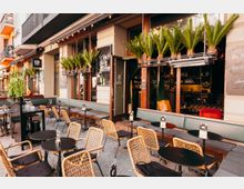 Außenbereich eines Straßencafés mit runden schwarzen Tischen, Korbstühlen und gepolsterten Sitzbänken entlang der Fassade. Große Fensterfronten mit offenen Türen, Pflanzkästen mit hohen Palmen und eine Lichterkette unter dem Vordach prägen die Szene.