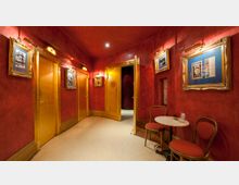 Ein schmaler Innenraum mit roten Wänden, goldenen Türen und gerahmten Bildern an den Wänden. Im Vordergrund steht ein kleiner Tisch mit zwei gepolsterten Stühlen, während schwaches Deckenlicht den Raum beleuchtet.